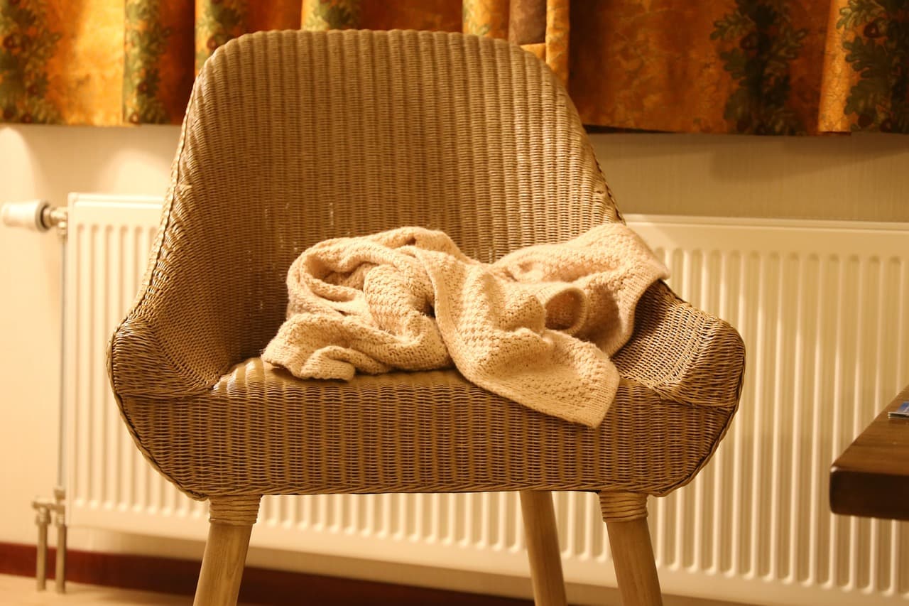 A white panel radiator under a window beside a wicker chair with a towel